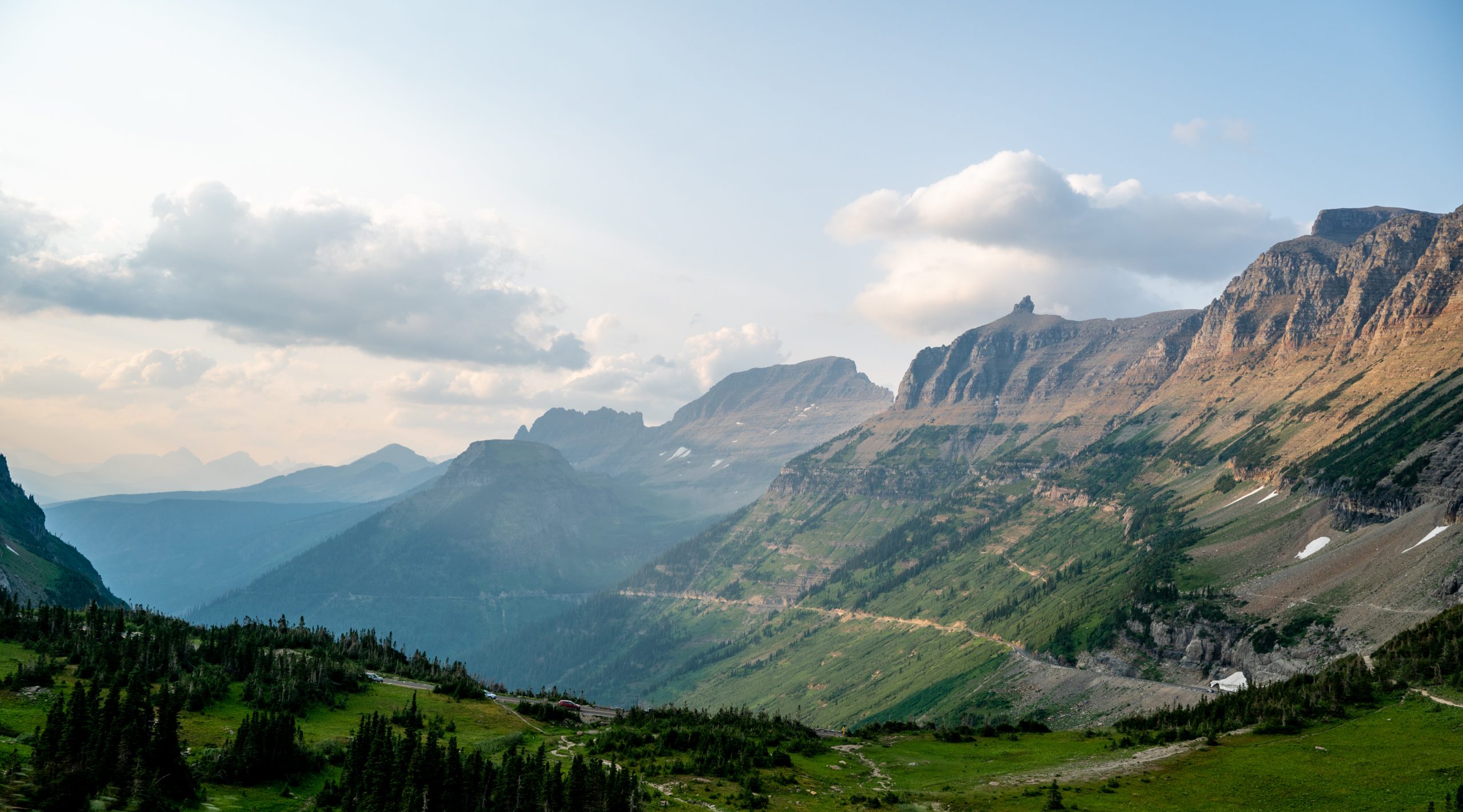 Mountain landscape view of the Garden Wall in Glacier National Park, Montana, with layered mountain ridges, green alpine meadows, and a hazy sky scattered with soft clouds in the evening light.