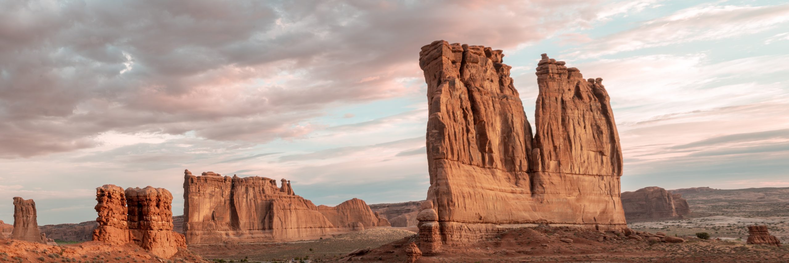 A panoramic view of the Three Gossips rock formation and Courthouse Towers glowing in warm sunrise light beneath a dramatic, cloud-filled sky in Arches National Park, Utah.