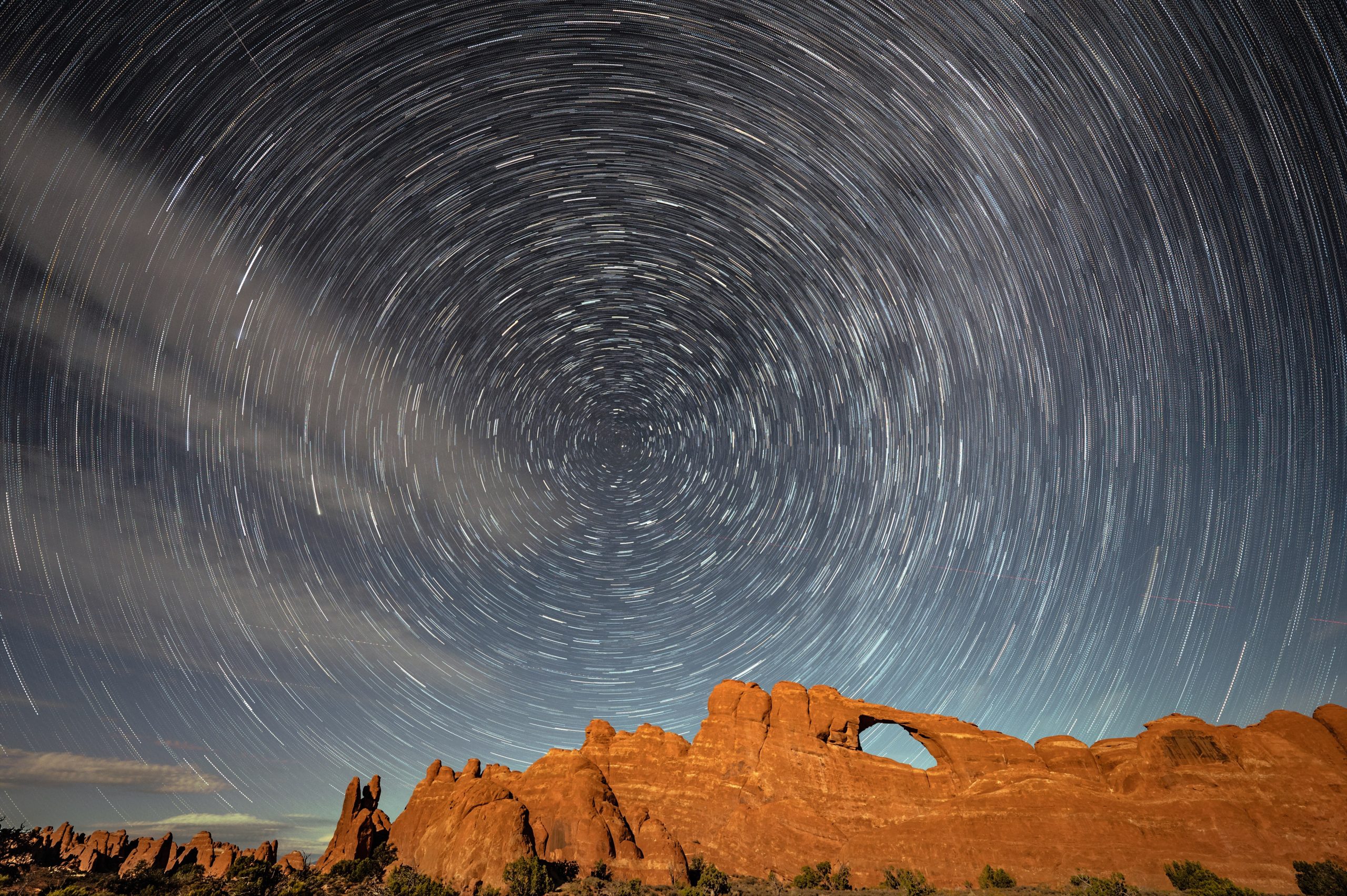 Long exposure star trail photograph over sandstone formations in Arches National Park, Utah, with spiraling night sky stars and illuminated red rock landscape in the foreground.